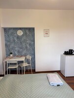 Una habitación con una cama, una mesa pequeña con sillas, un reloj, un cuadro enmarcado y un refrigerador.