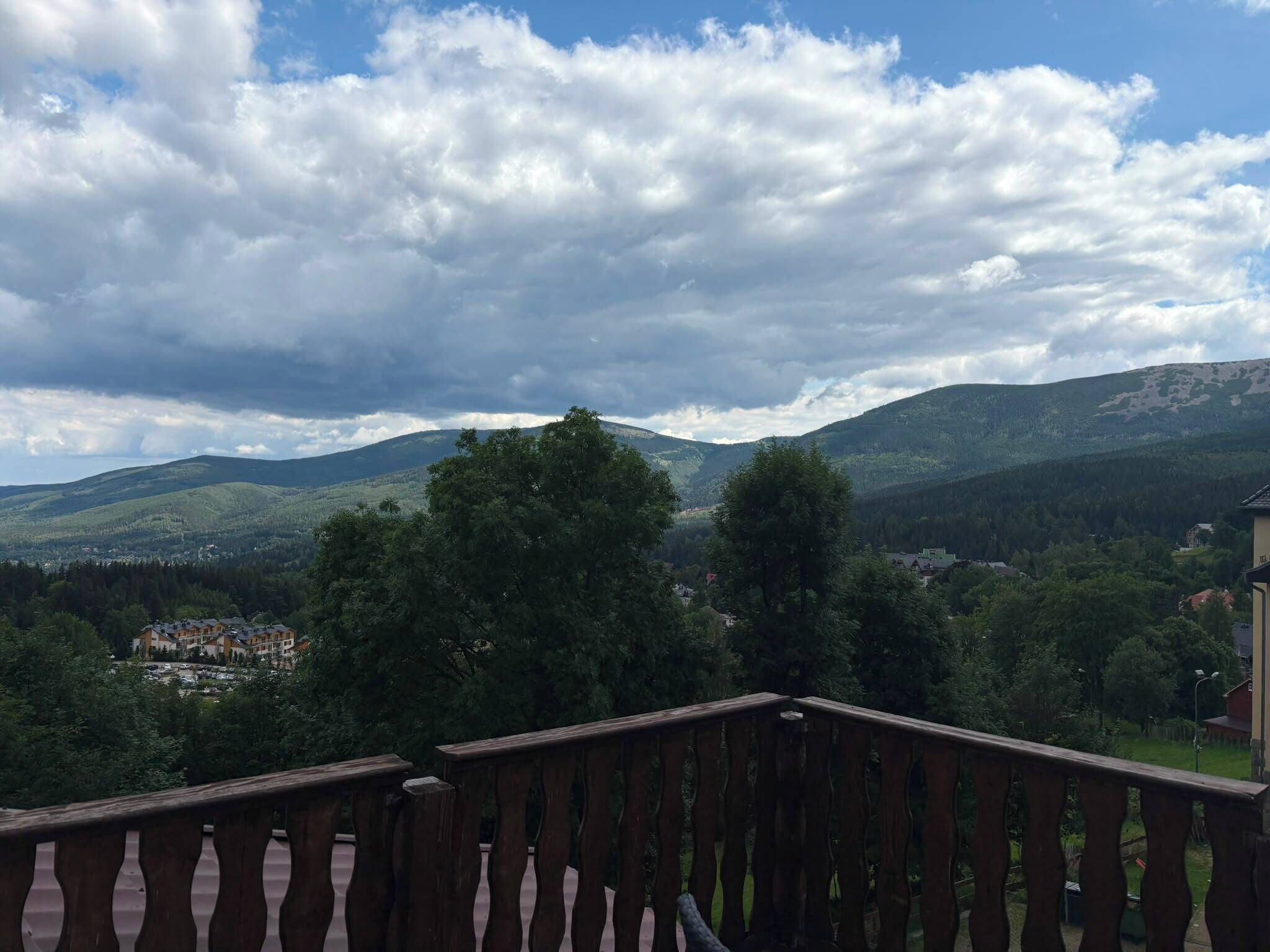Uitzicht vanaf een balkon met een hekwerk, over een vallei met bomen en een bergketen in de verte.
