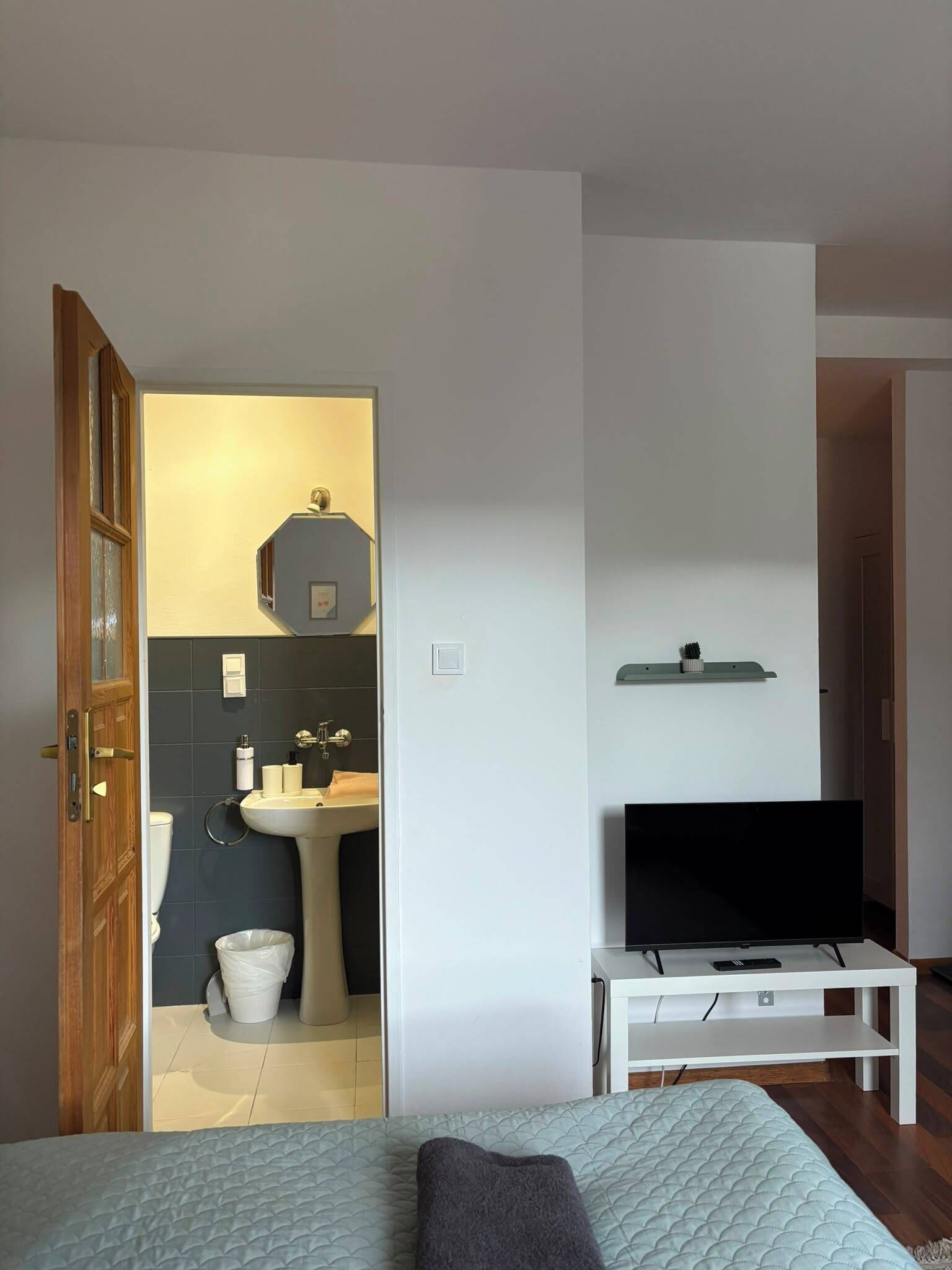 Un dormitorio con cama, televisor sobre un mueble, baño con lavamanos e inodoro, y una puerta que da a otra habitación.