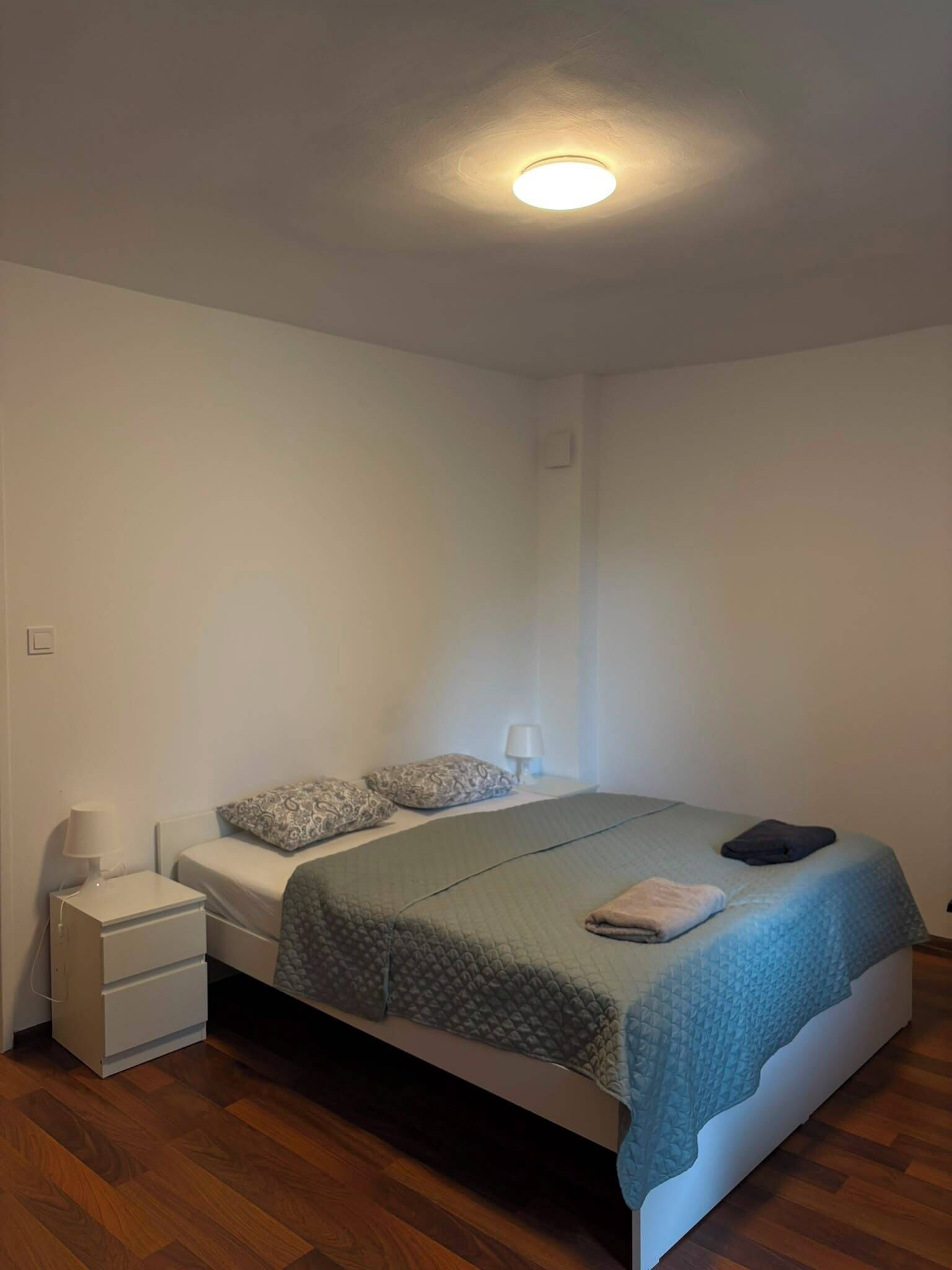 Habitación individual con cama, suelo de madera, mesita de noche con lámpara y ropa de cama con colcha y almohadas.