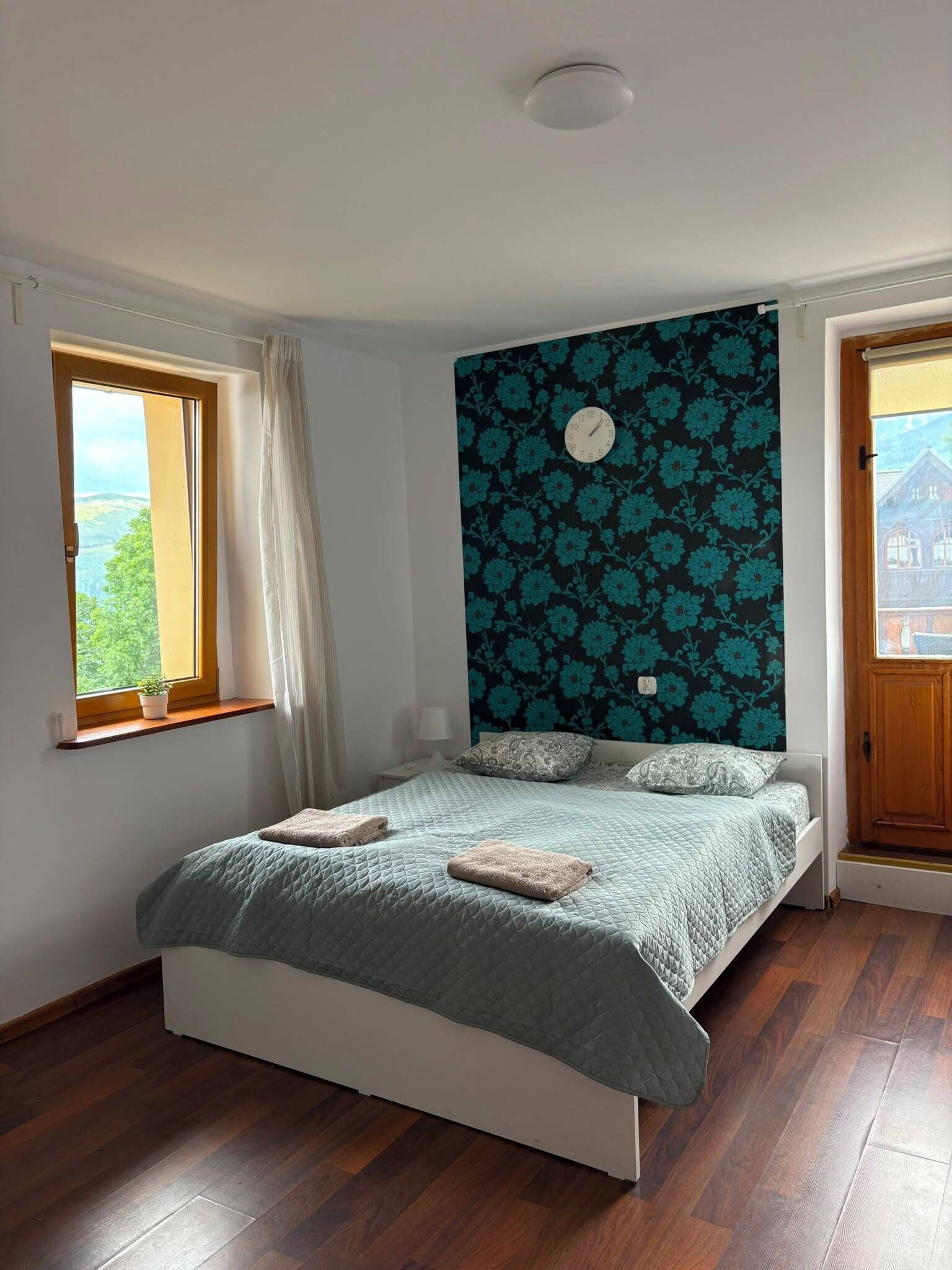 A bedroom with a bed, a window, a clock, and a view of the outside.