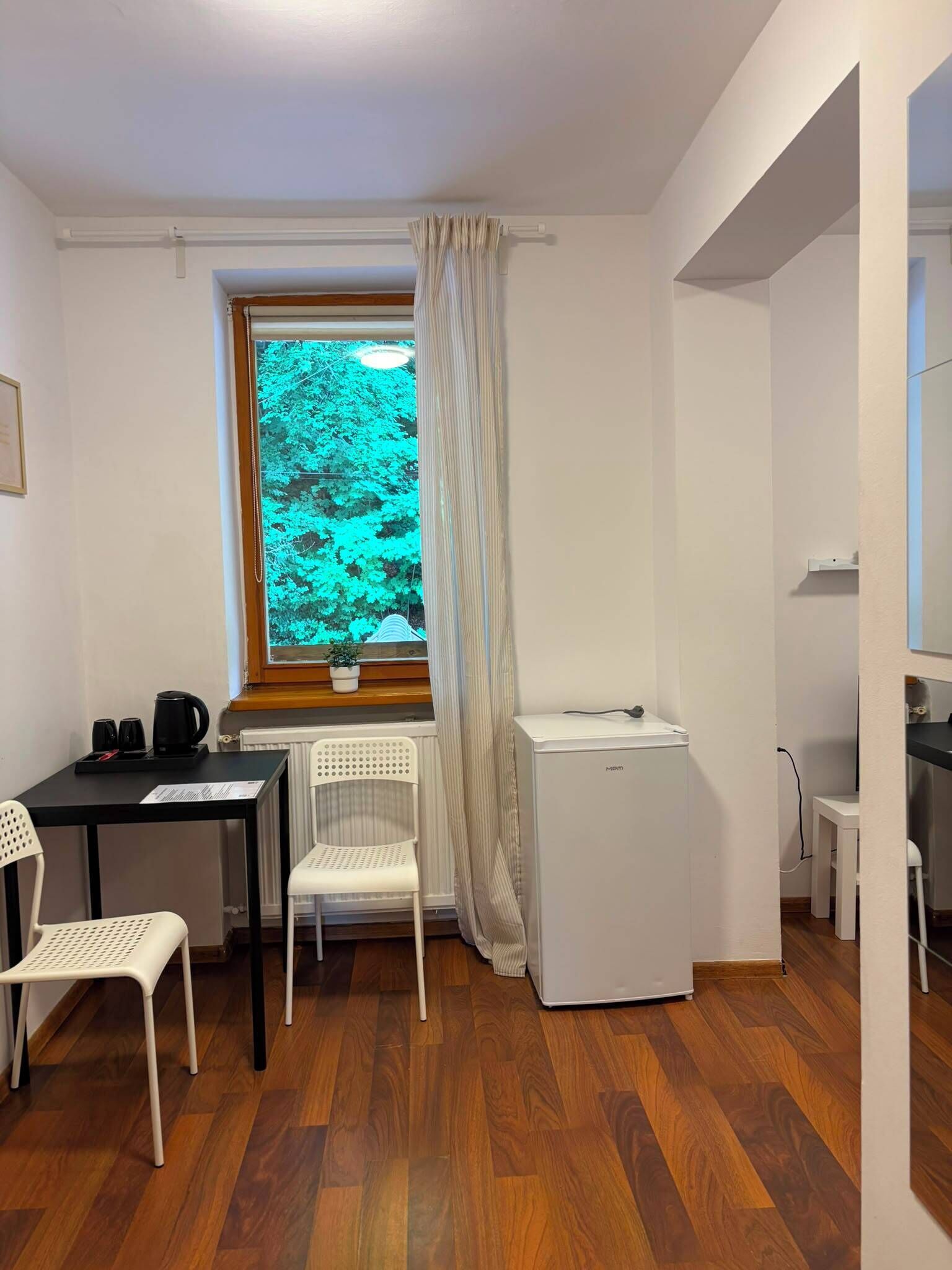 A small, compact room with a wooden floor, a white chair, a small table with a kettle, a window with a view of trees, and a white appliance.