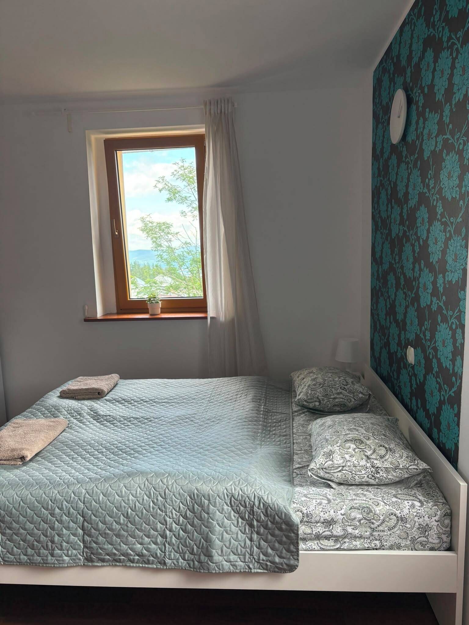 A single bed with a quilted headboard, a window with a view of greenery, and a wall with a floral pattern.