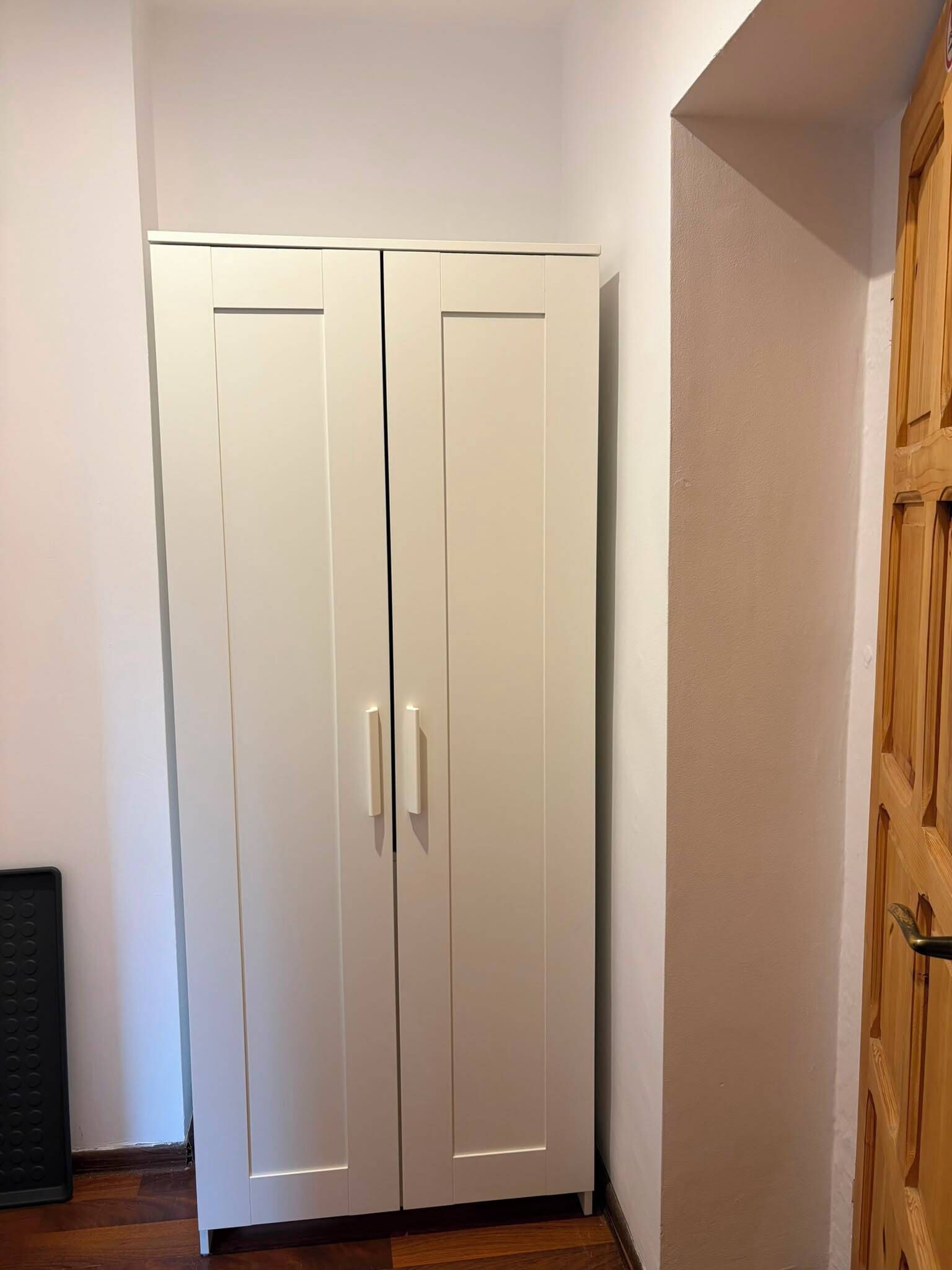Un armario blanco con dos puertas y un tirador, ubicado en una habitación con suelo de madera y marco de puerta de madera.