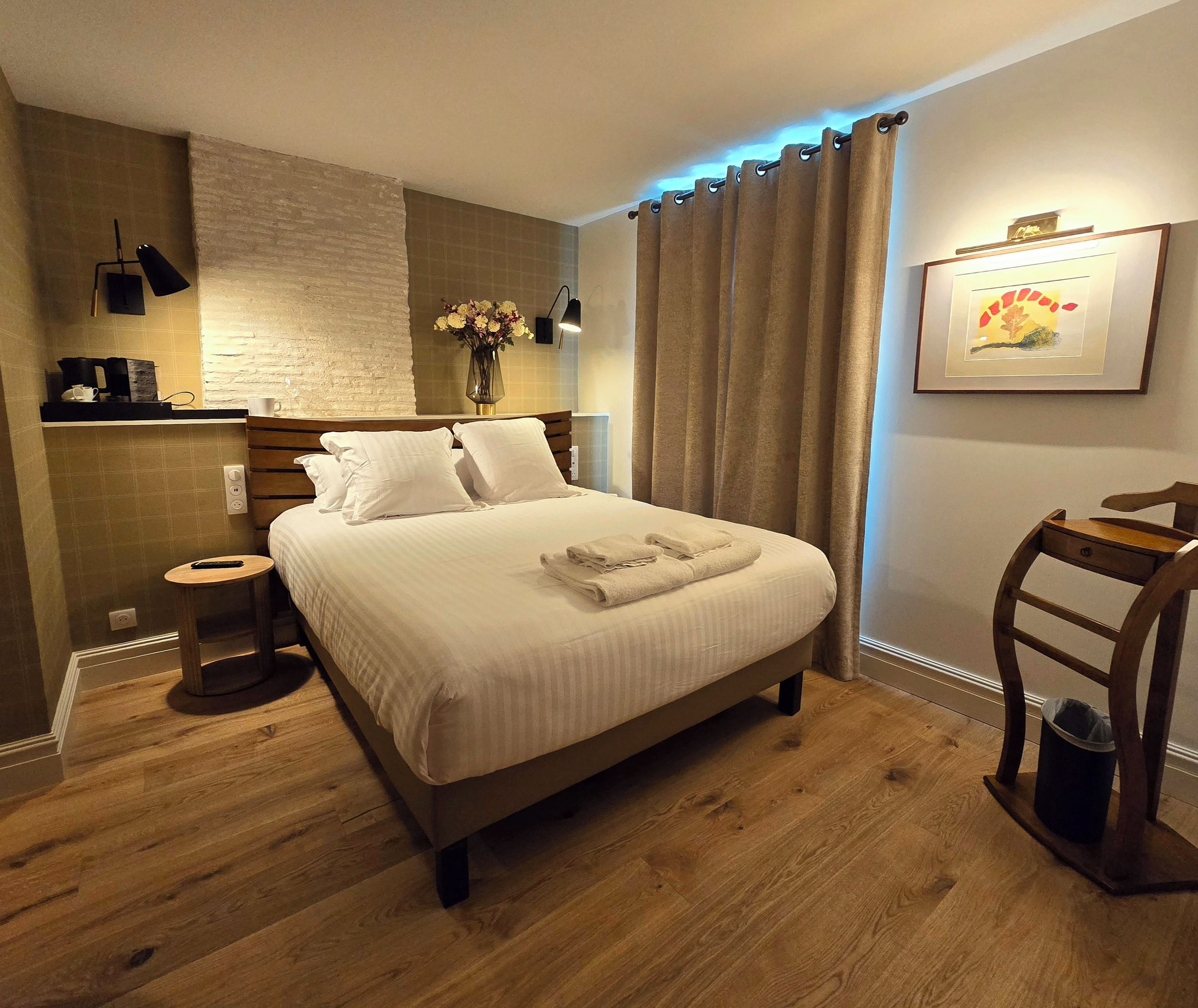 Un lit bien fait, avec du linge de lit et des serviettes blanches, une table de chevet avec une télécommande, une lampe fixée au mur et un tableau encadré accroché au mur.