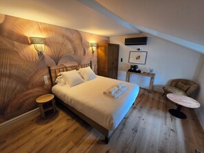 A bedroom with a bed, bedside tables, a chair, a small table, a wall-mounted TV, and a wooden floor. - Le Groom Hôtel (Laprade)