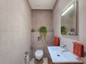 Ein modernes Badezimmer mit einem weißen Waschbecken, einer Toilette und einer Dusche. Über dem Waschbecken befindet sich ein Spiegel und an der Wand ein Topf mit einer Pflanze.