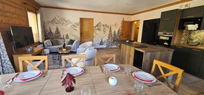A dining area with a wooden table set for four, a sofa, and a kitchen area with a refrigerator and stove. - L'Ecrin Alpin- Votre parenthèse alpine ski au pied aux Menuires- 4 chambres (Les Belleville)