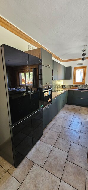 A modern kitchen with a built-in oven, a refrigerator, and a dining area with a table and chairs. - L'Ecrin Alpin- Votre parenthèse alpine ski au pied aux Menuires- 4 chambres (Les Belleville)