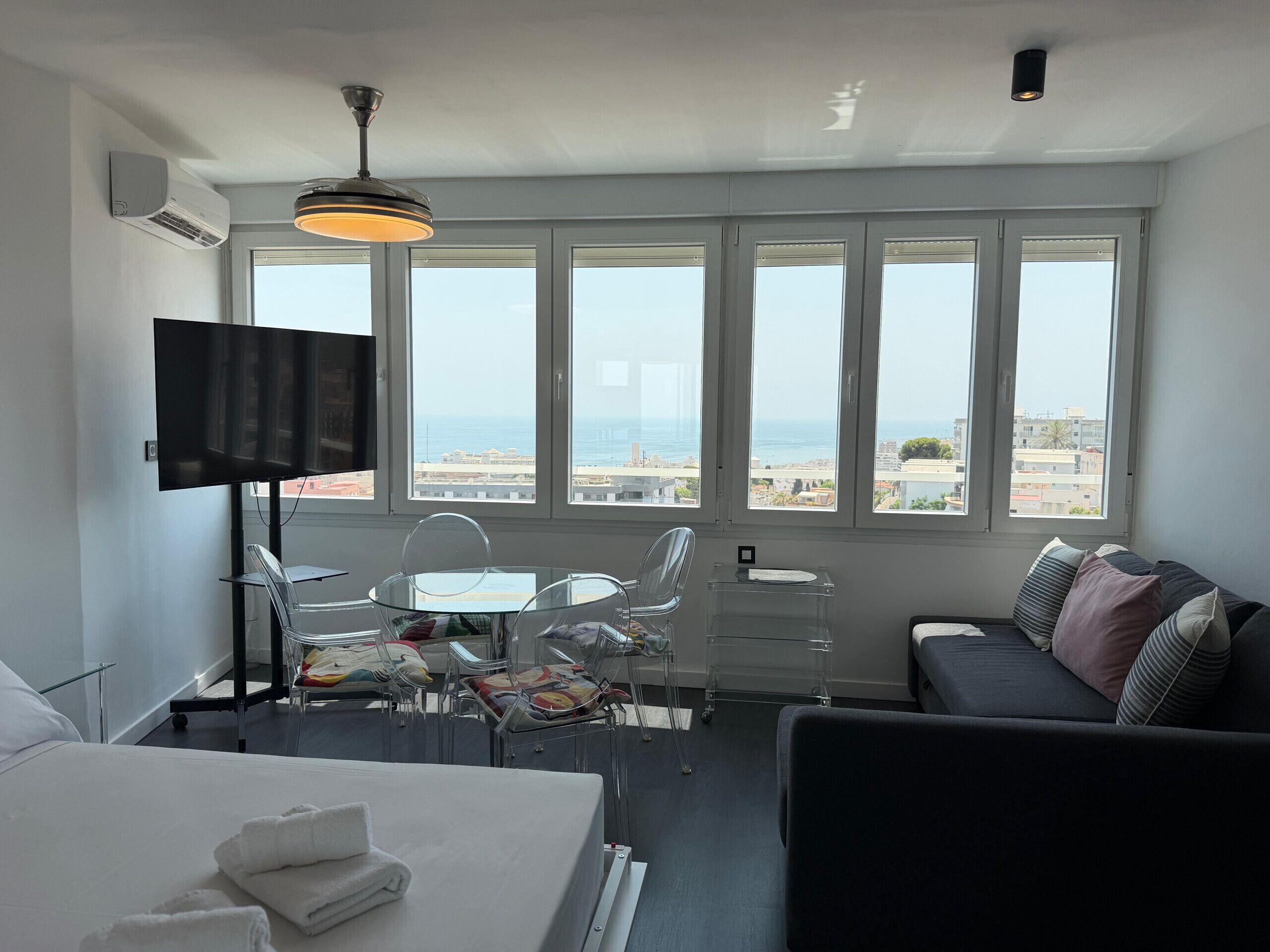 A hotel room with a bed, a TV, a desk with a chair, a sofa, and large windows with a view of the sea.