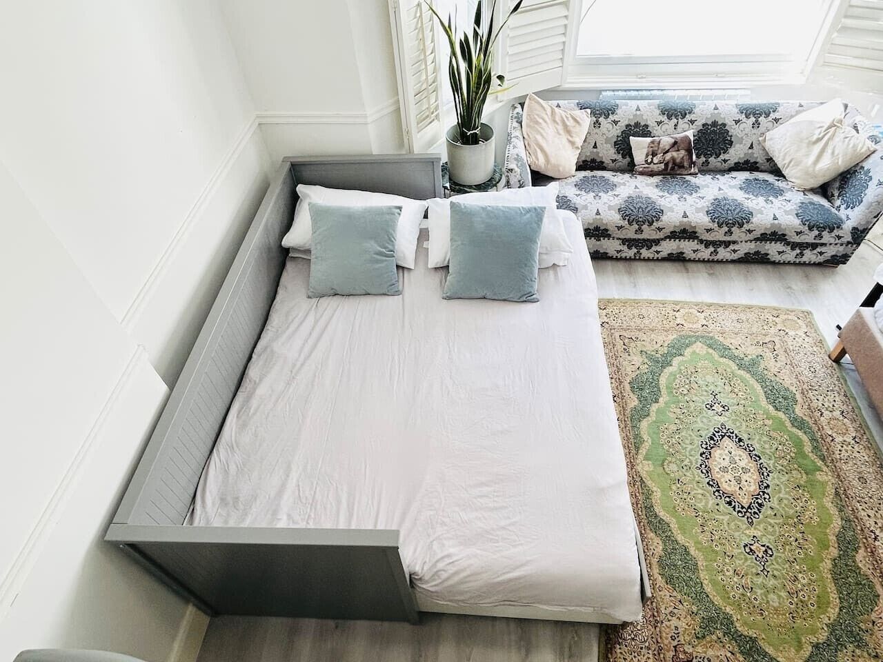 Una cama con ropa de cama blanca y dos almohadas, un sofá estampado con cojines, una planta en maceta y un tapete decorativo.