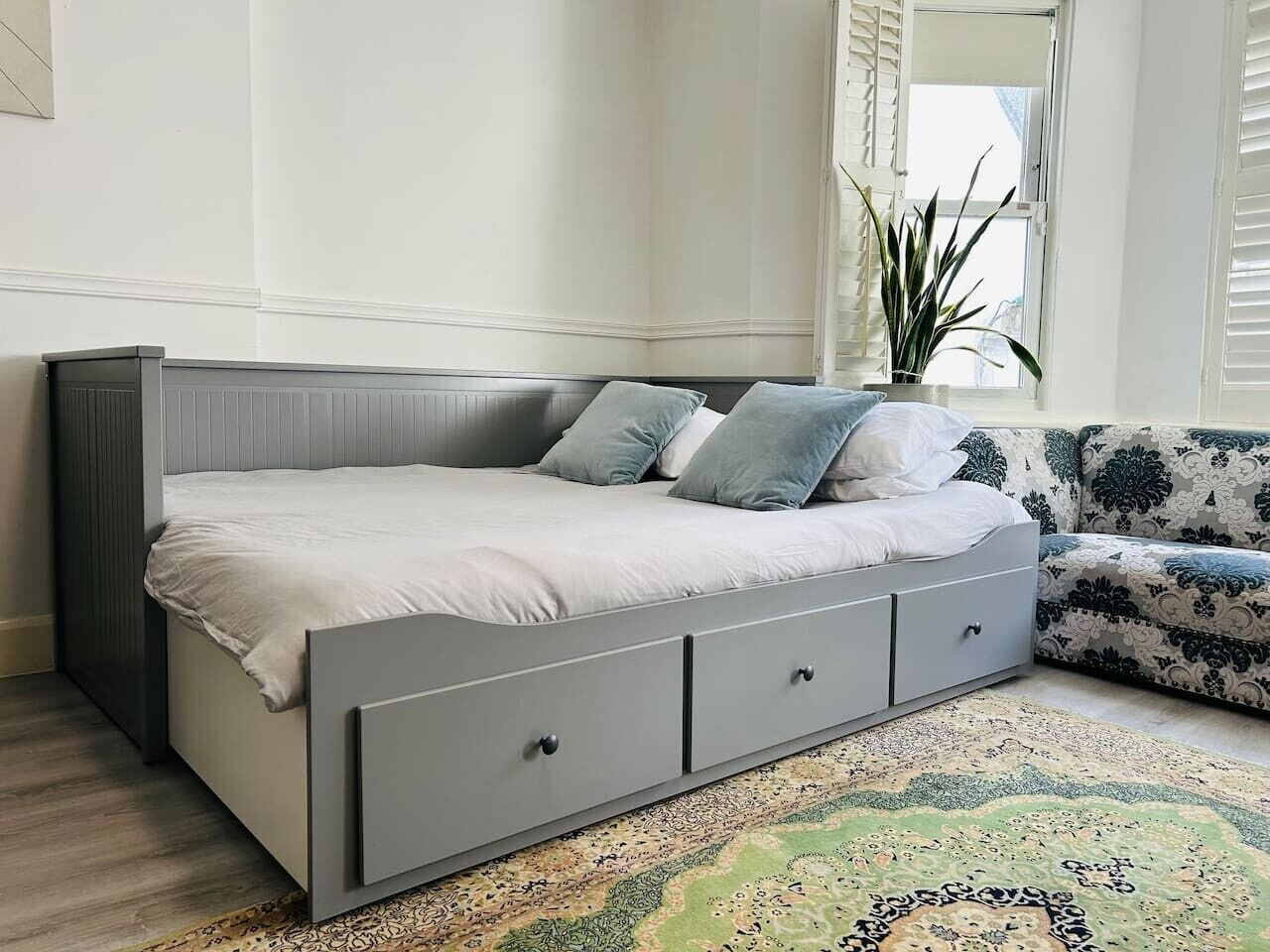 Quarto moderno com uma cama cinzenta, um tapete estampado e uma janela com persianas.
