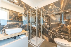 A modern bathroom with a glass shower enclosure, a white sink, and a marble-tiled wall. - Suite with a terrace overlooking the lake | Lagorá – C7 (San Giuliano Milanese)