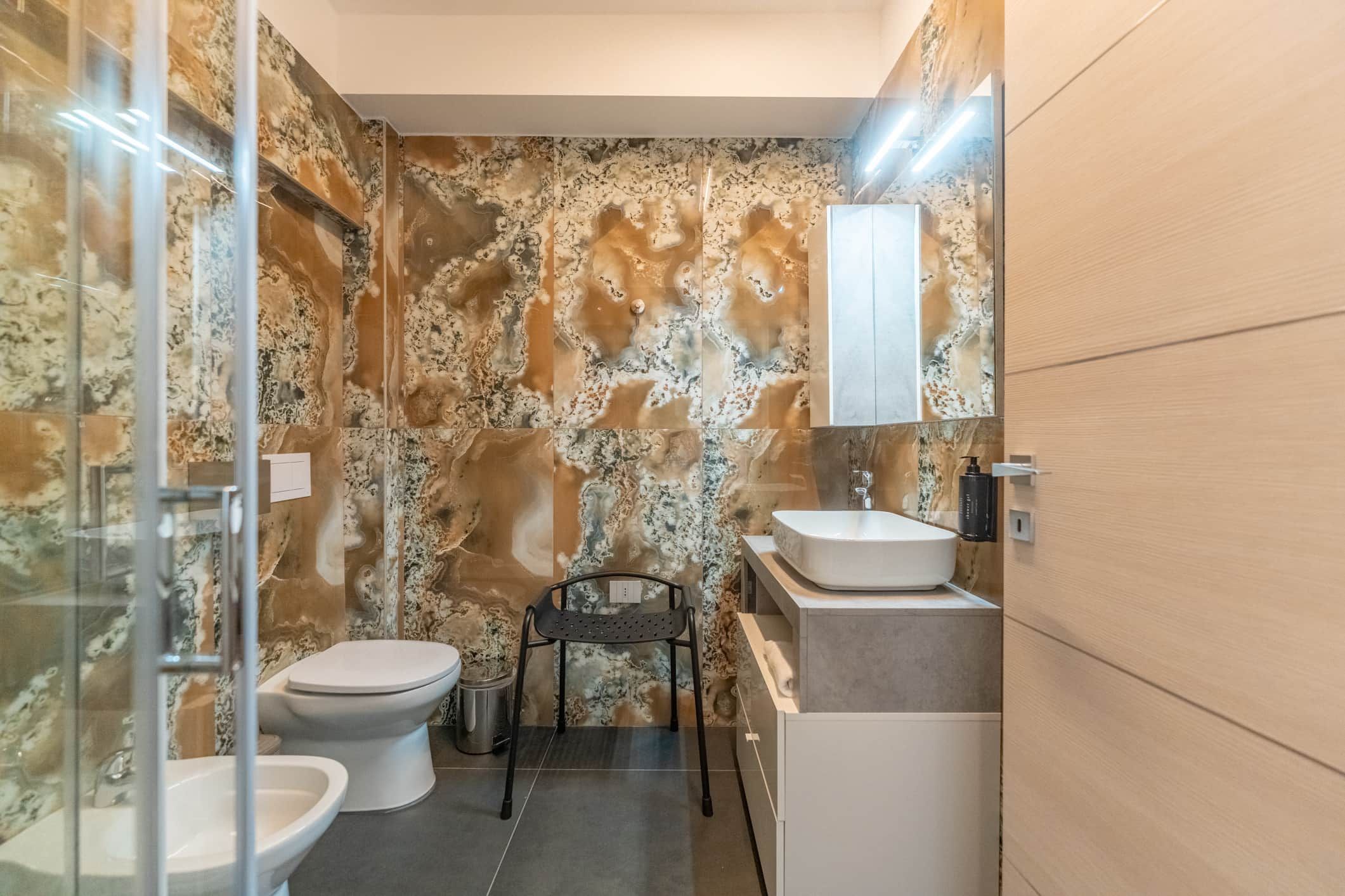 Een badkamer met een marmeren wand, een toilet, een bidet, een wastafel en een stoel.