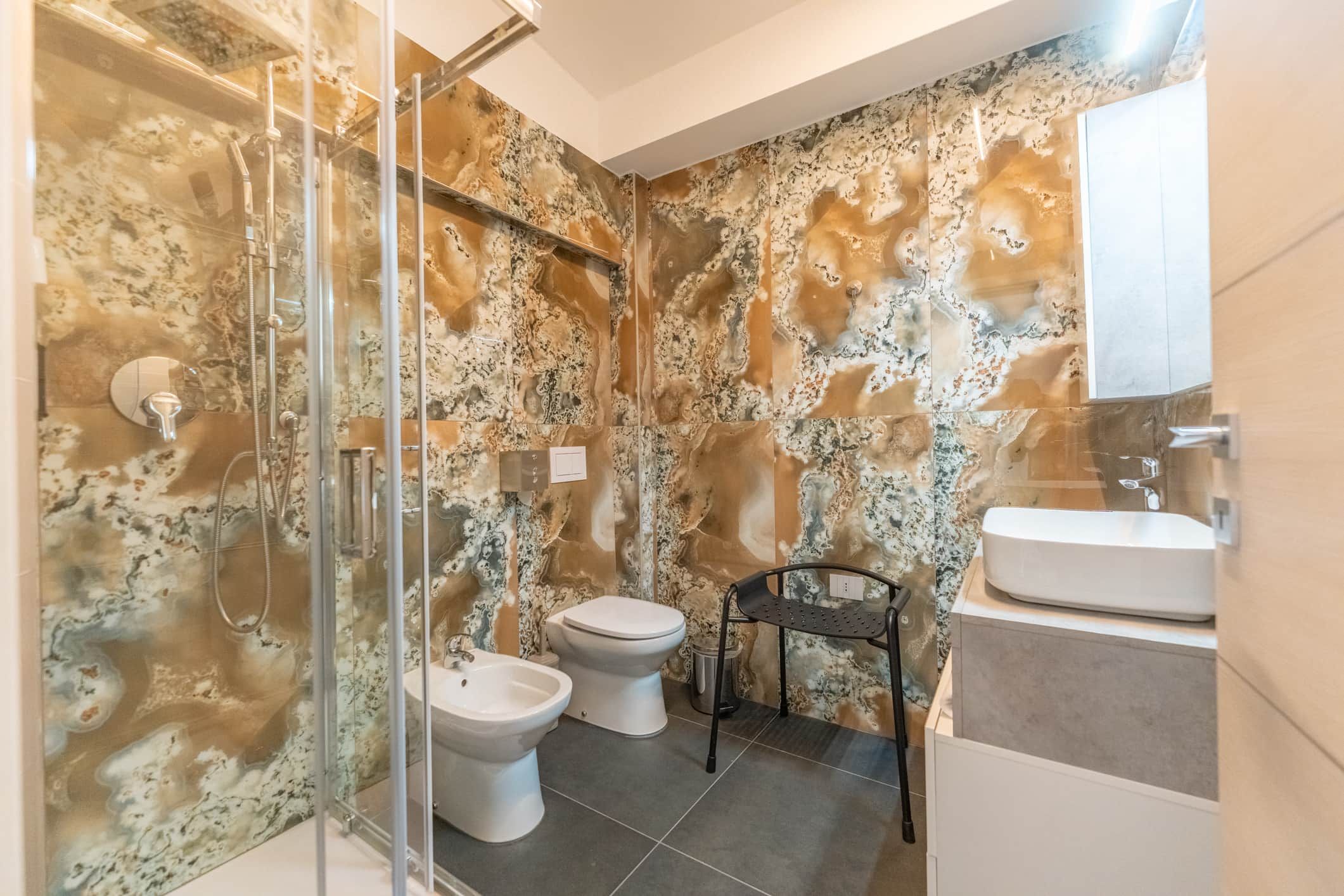 A bathroom with a glass-enclosed shower, two toilets, a sink, and a marble-patterned wall.