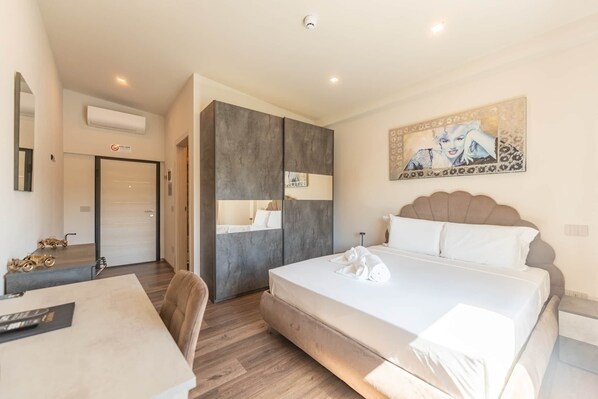 A hotel room with a bed, a desk, a chair, a wardrobe, and a painting on the wall.