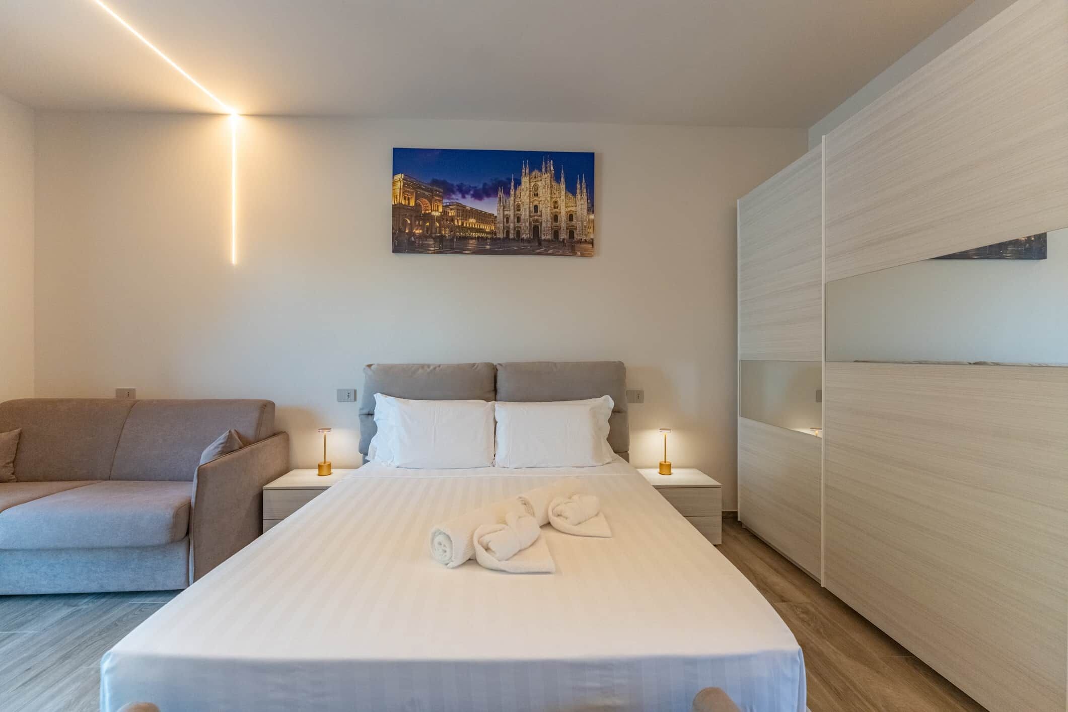 A hotel room with a bed, a sofa, a nightstand with a lamp, and a painting on the wall.