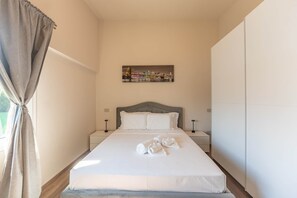 Una habitación de hotel con cama, dos mesitas de noche, una lámpara de noche, una ventana con cortinas y un cuadro de paisaje urbano en la pared.