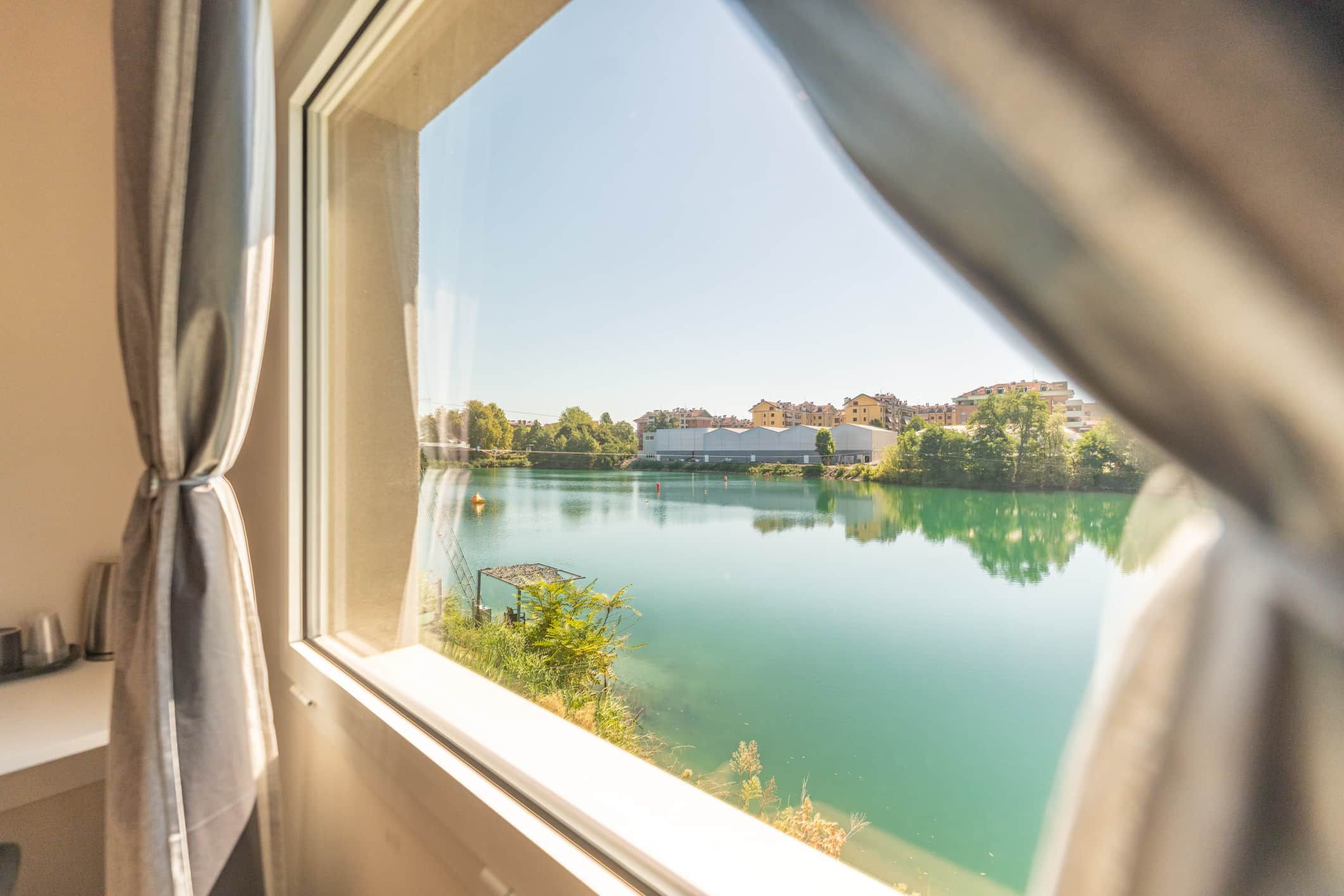 Una vista attraverso una finestra con tende, che mostra un lago tranquillo, edifici sullo sfondo e vegetazione lungo la riva.