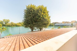 A rooftop terrace with a view of a calm body of water, a tree, and buildings in the background. - Suite with a terrace overlooking the lake | Lagorá – C6 (San Giuliano Milanese)