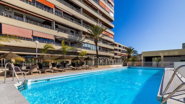 Une piscine entourée de chaises longues et de parasols, elle-même bordée d’un immeuble à plusieurs étages doté de balcons.