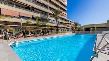 A swimming pool with lounge chairs and umbrellas, surrounded by a multi-story building with balconies.