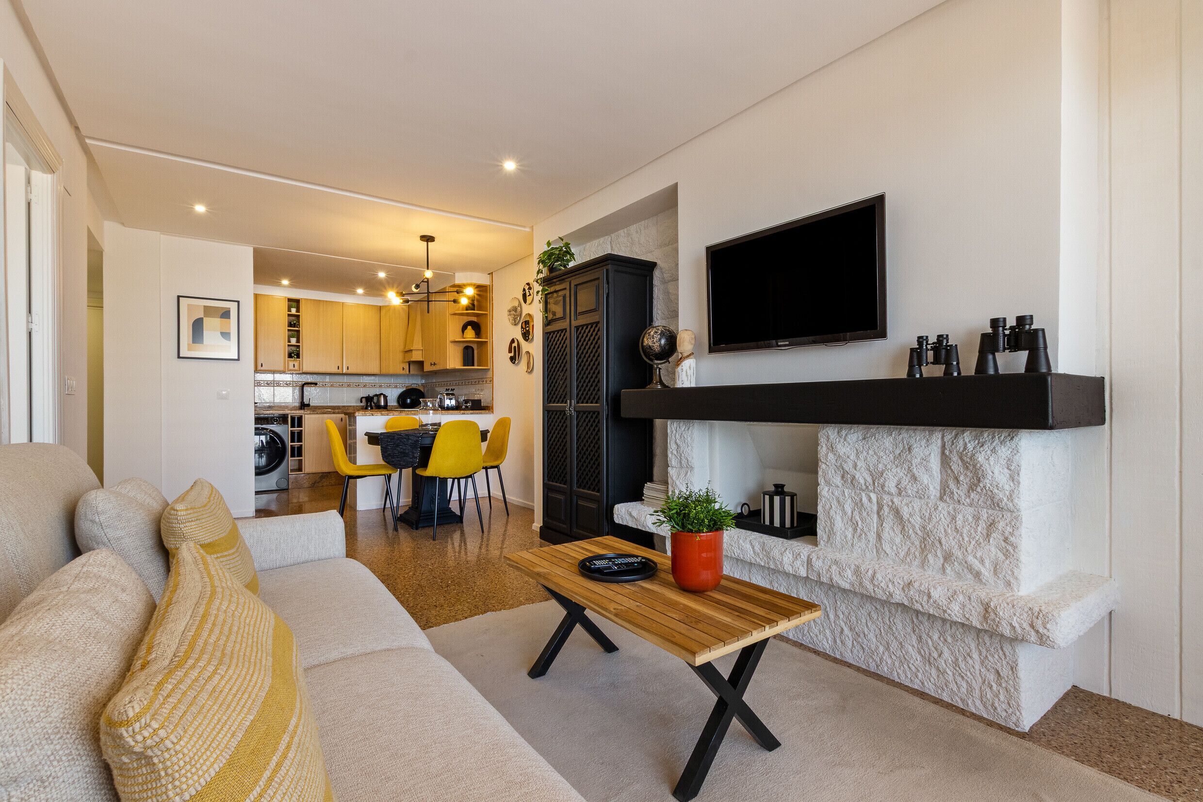 Un salon moderne avec un canapé, un coin repas avec des chaises jaunes, une cuisine en arrière-plan et un téléviseur fixé au mur.