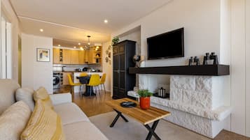 A modern living room with a sofa, a dining area with yellow chairs, a kitchen in the background, and a mounted TV on the wall.