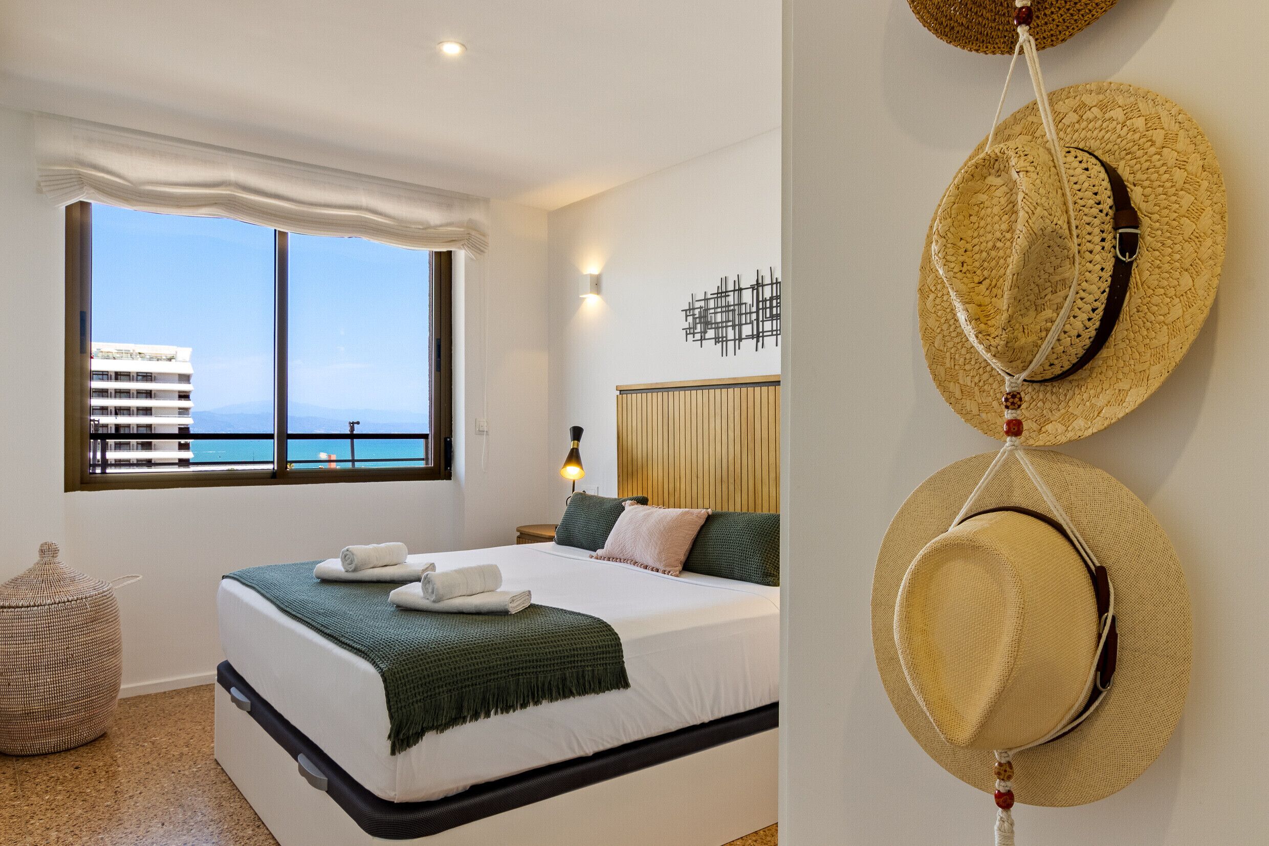 A hotel room with a large window, a bed with a green throw, and two hats hanging on the wall.