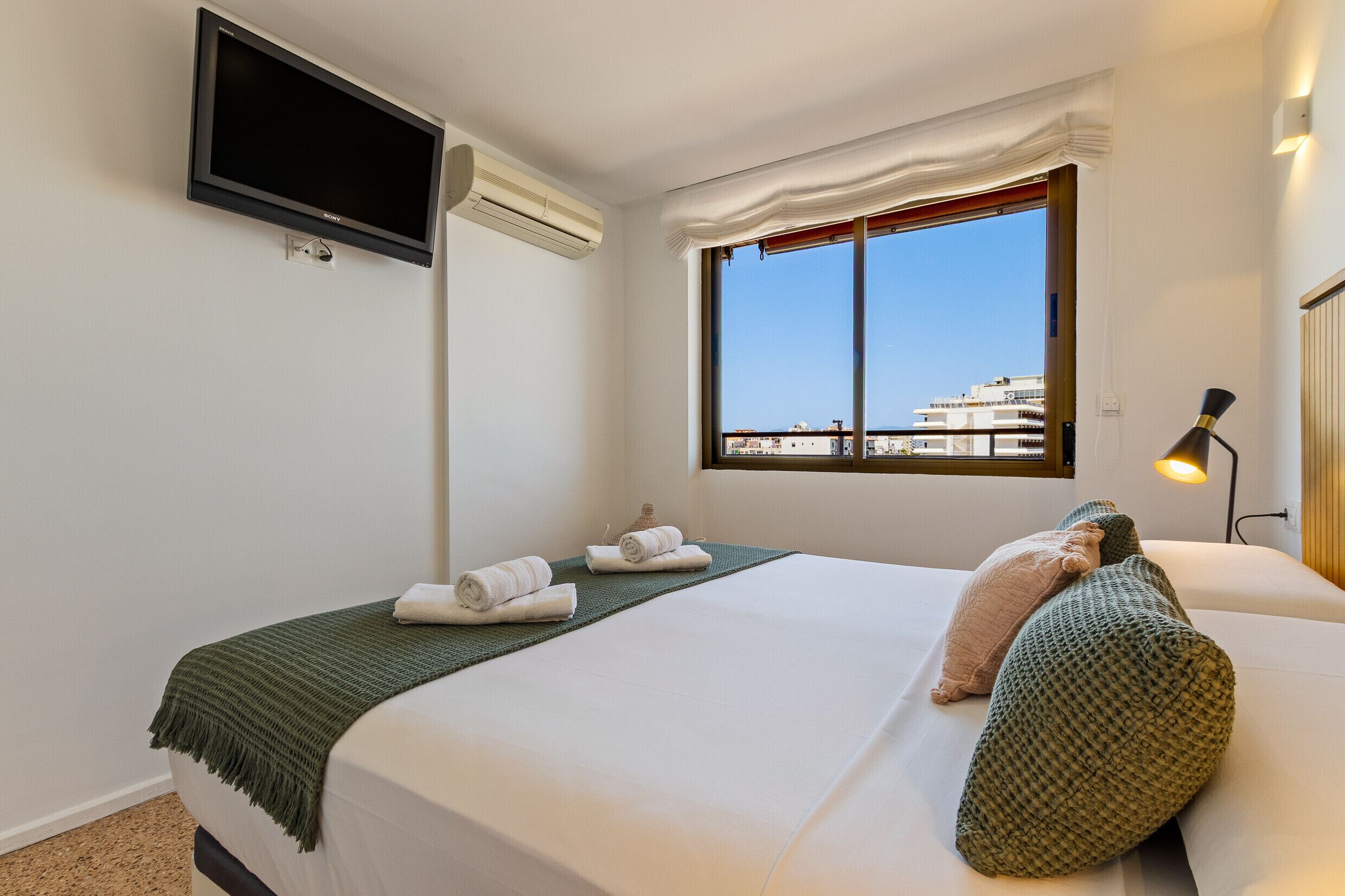 A hotel room with a bed, a flat-screen TV mounted on the wall, an air conditioning unit, a window with a view of buildings, and a bedside lamp.