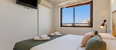 A hotel room with a bed, a flat-screen TV mounted on the wall, an air conditioning unit, a window with a view of buildings, and a bedside lamp.