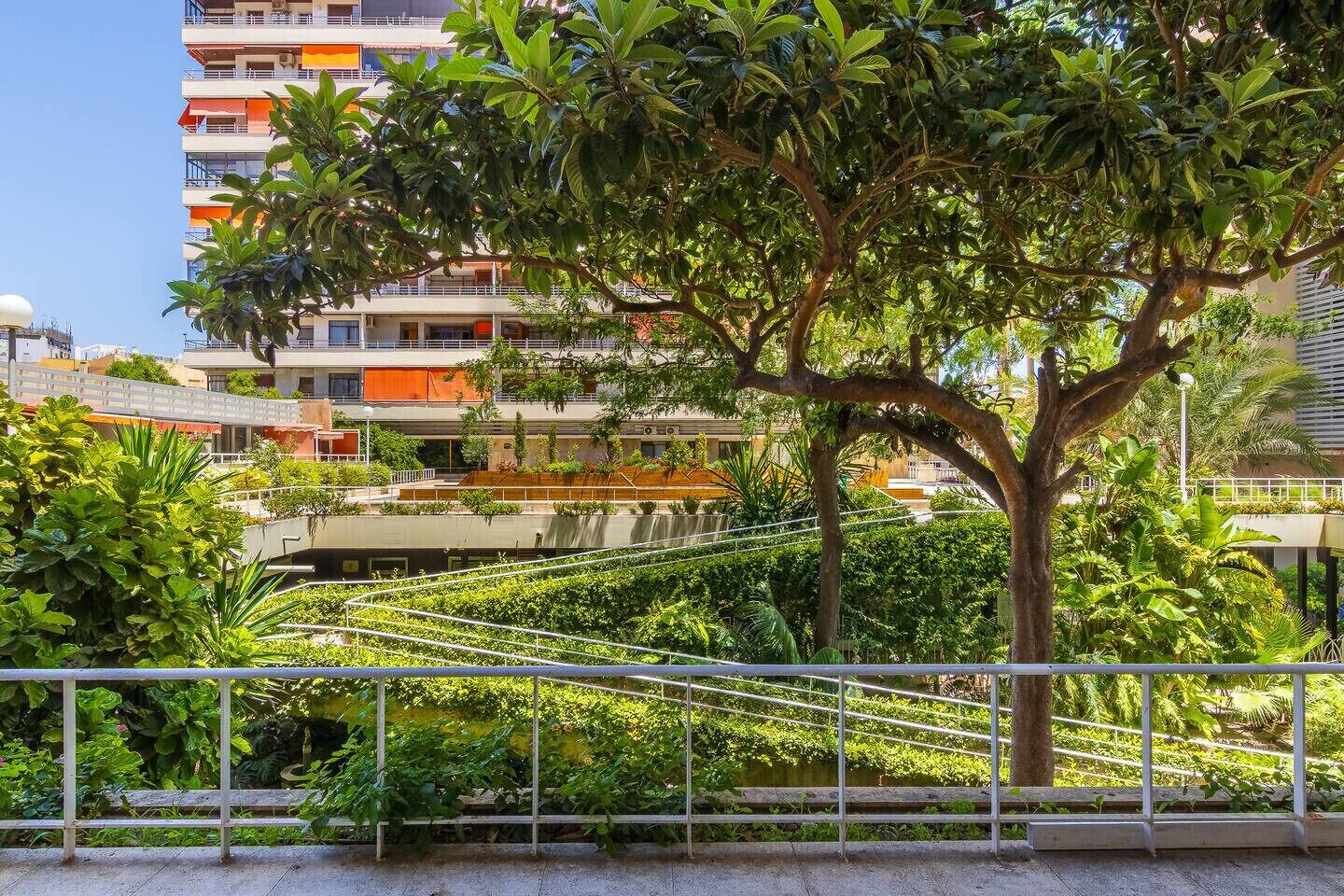 A multi-story building with balconies, surrounded by lush greenery and a metal fence.
