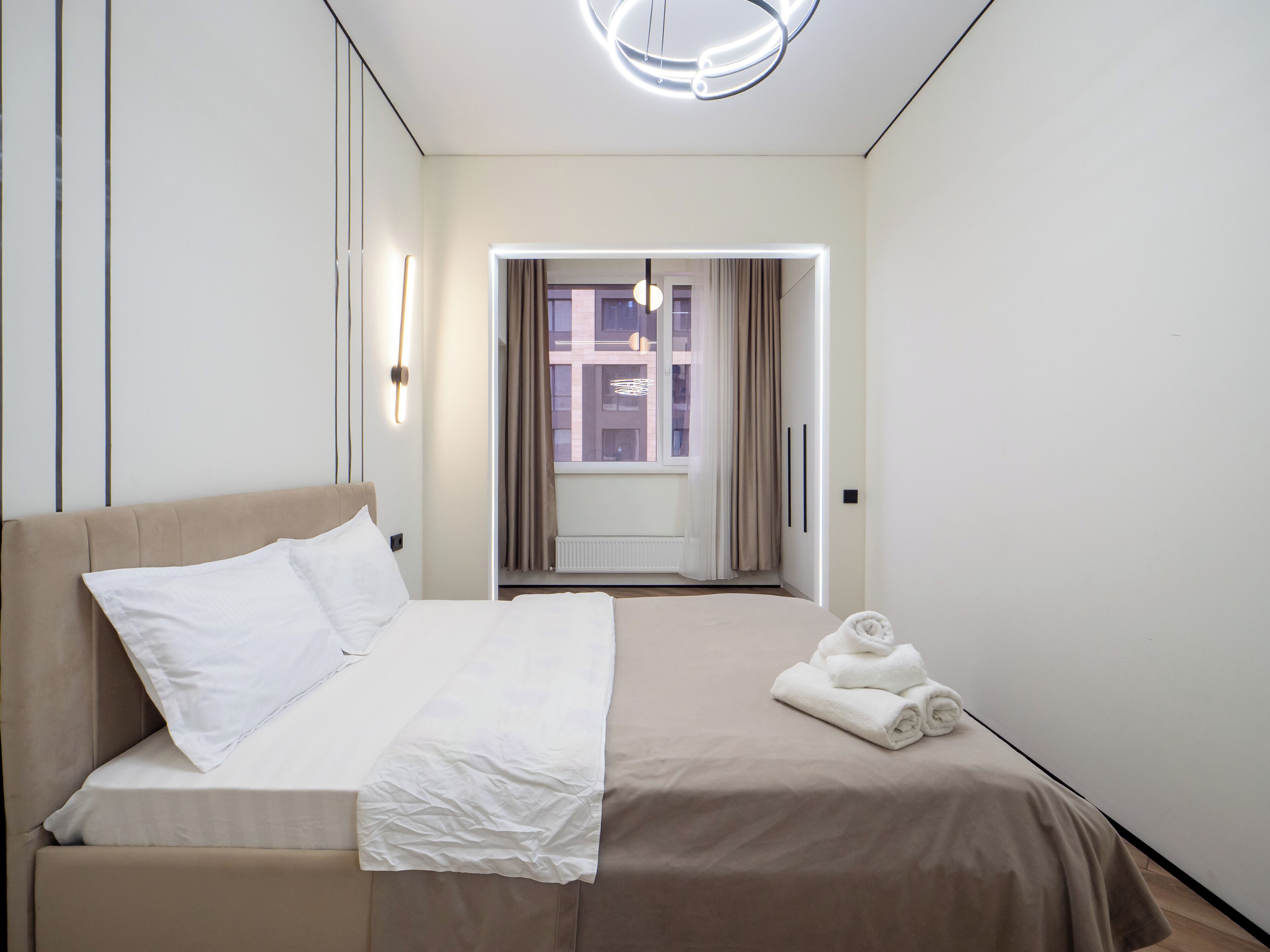A modern hotel room with a bed, a view through a window, and a neatly made bed with towels.