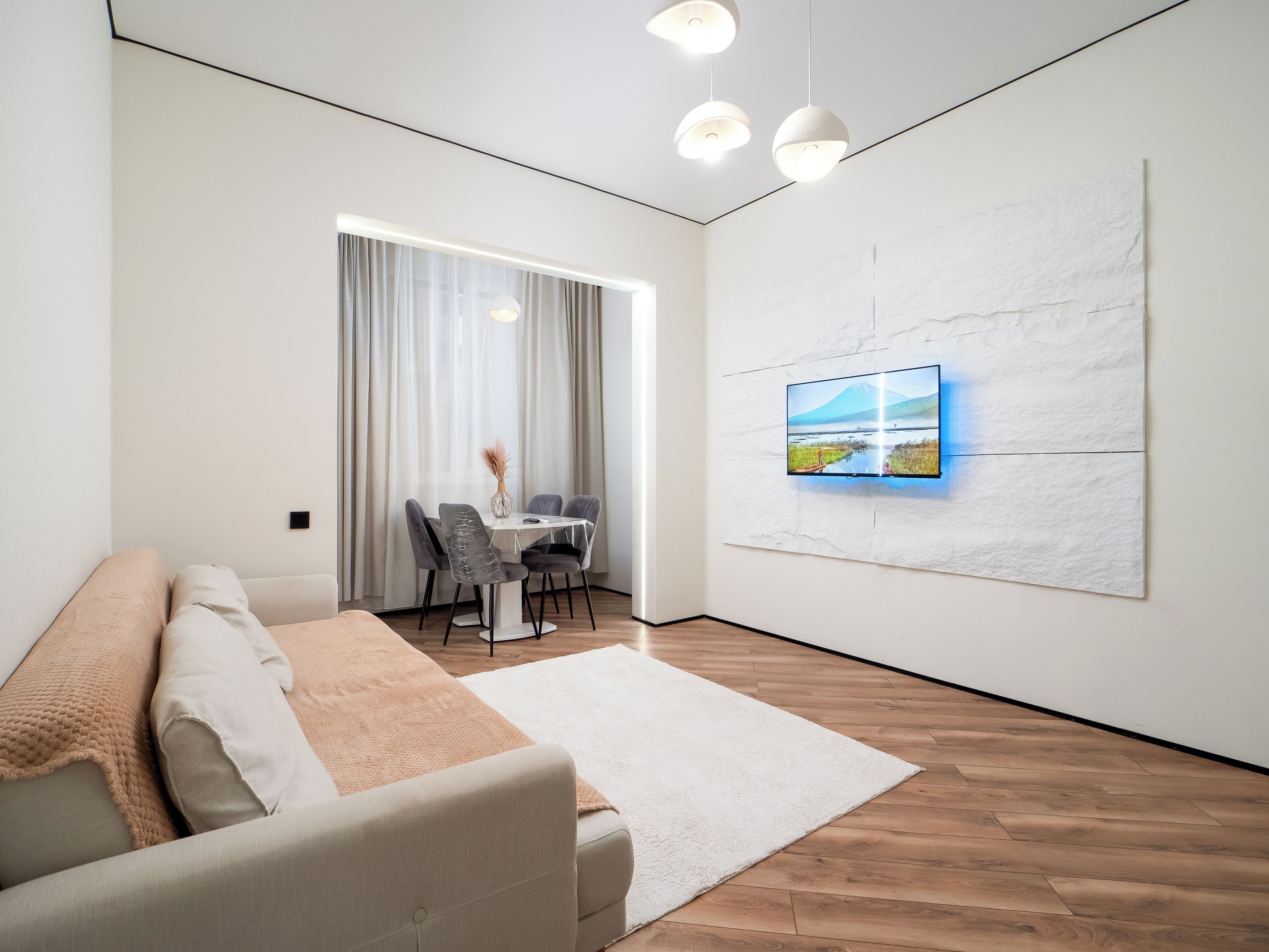 Una sala moderna con un sofá, una mesa de comedor con sillas, un televisor de pantalla plana en la pared y un amplio ventanal con cortinas.