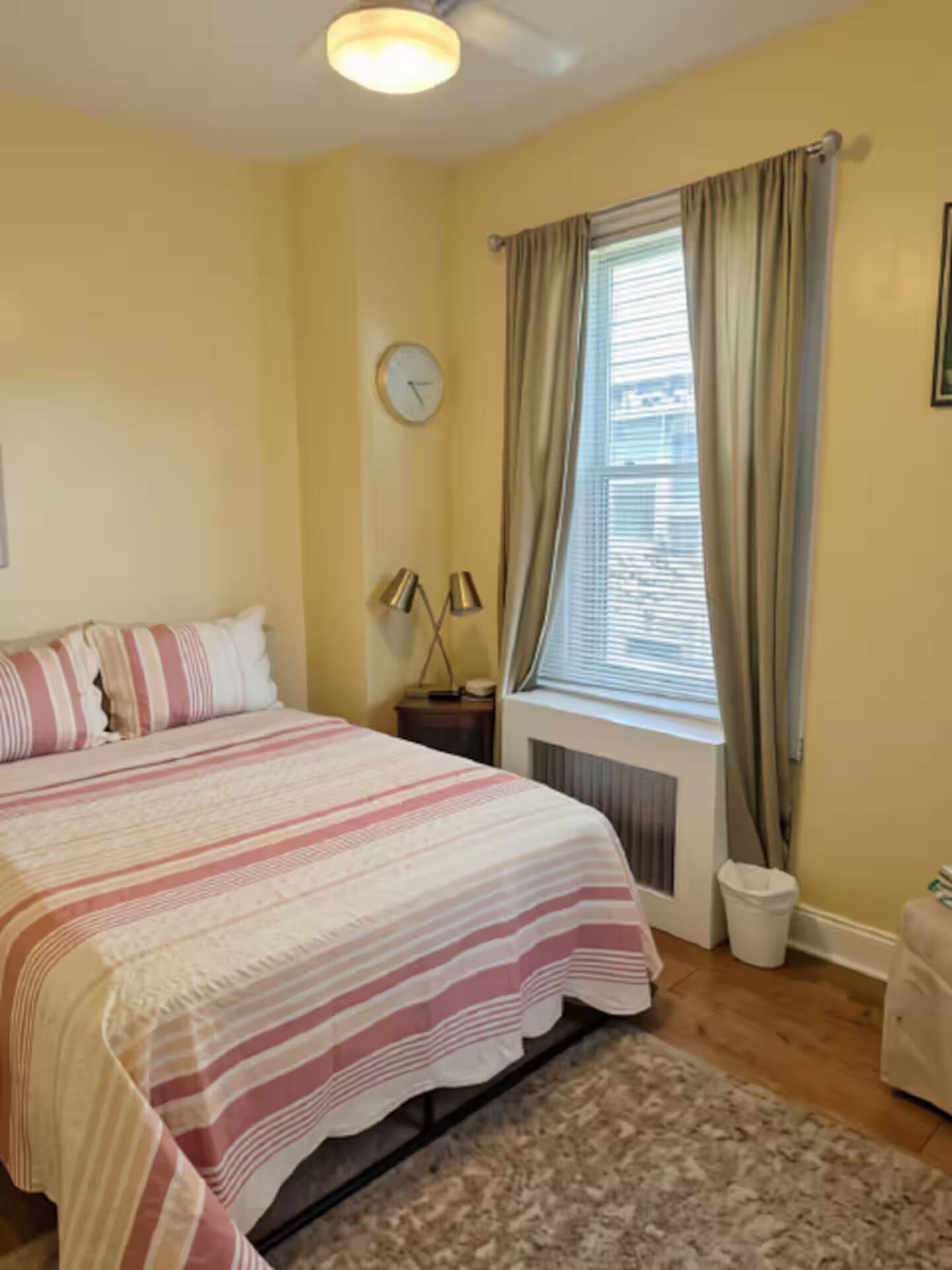 A hotel room with a bed, a nightstand, a lamp, a window with curtains, and a wall clock.