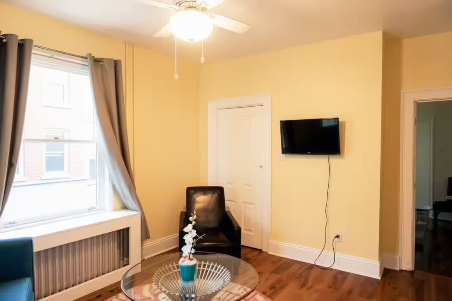 A room with a ceiling fan, a flat-screen TV mounted on the wall, a black leather chair, a glass table with a decorative item, and a window with curtains.