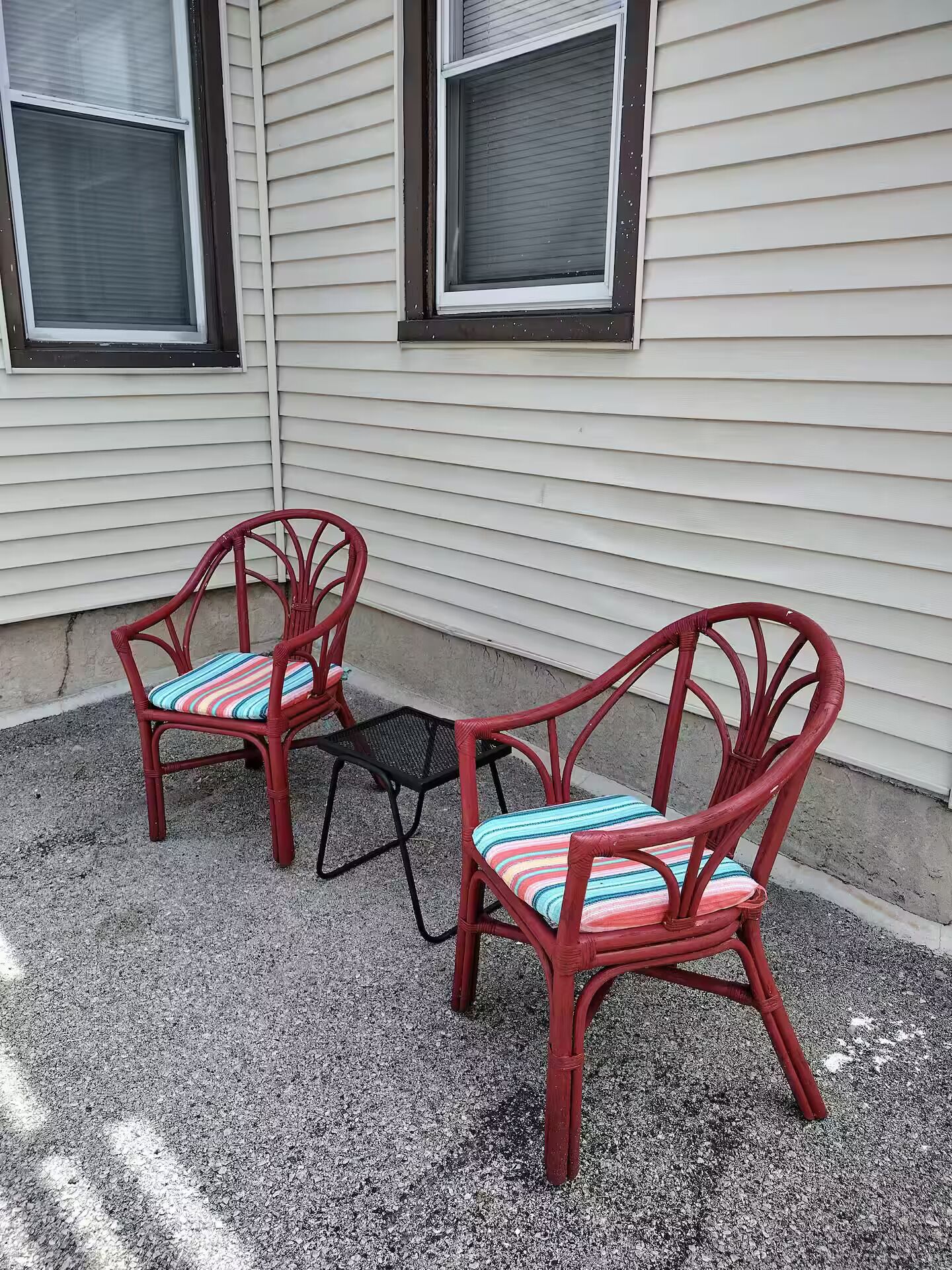 Two wicker chairs with striped cushions on a gravel surface, positioned in front of a house with white siding and two windows.