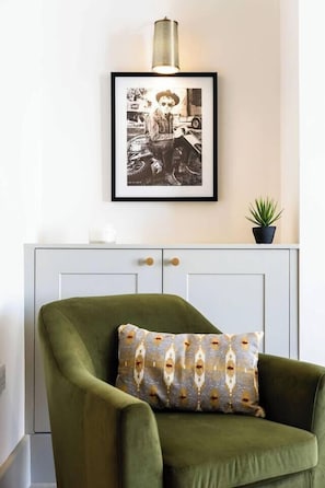 Ein Zimmer mit einem grünen Sessel, einem schwarzen und weißen Foto im Bilderrahmen an der Wand, einer kleinen Topfpflanze und einem Schrank mit zwei Türen.