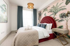 Ein Schlafzimmer mit rotem Kopfteil, einem Bett mit weißer Bettwäsche, einem Nachttisch und einer Wandmalerei von tropischen Pflanzen.
