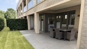 Ein modernes Gebäude mit einer Terrassenfläche, komplett mit Tisch und Stühlen, einem gepflegten Rasen und einem Heckenstreifen.