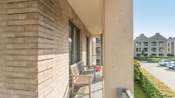 A balcony with a wooden bench, a glass door, and a view of a parking area and buildings in the background.