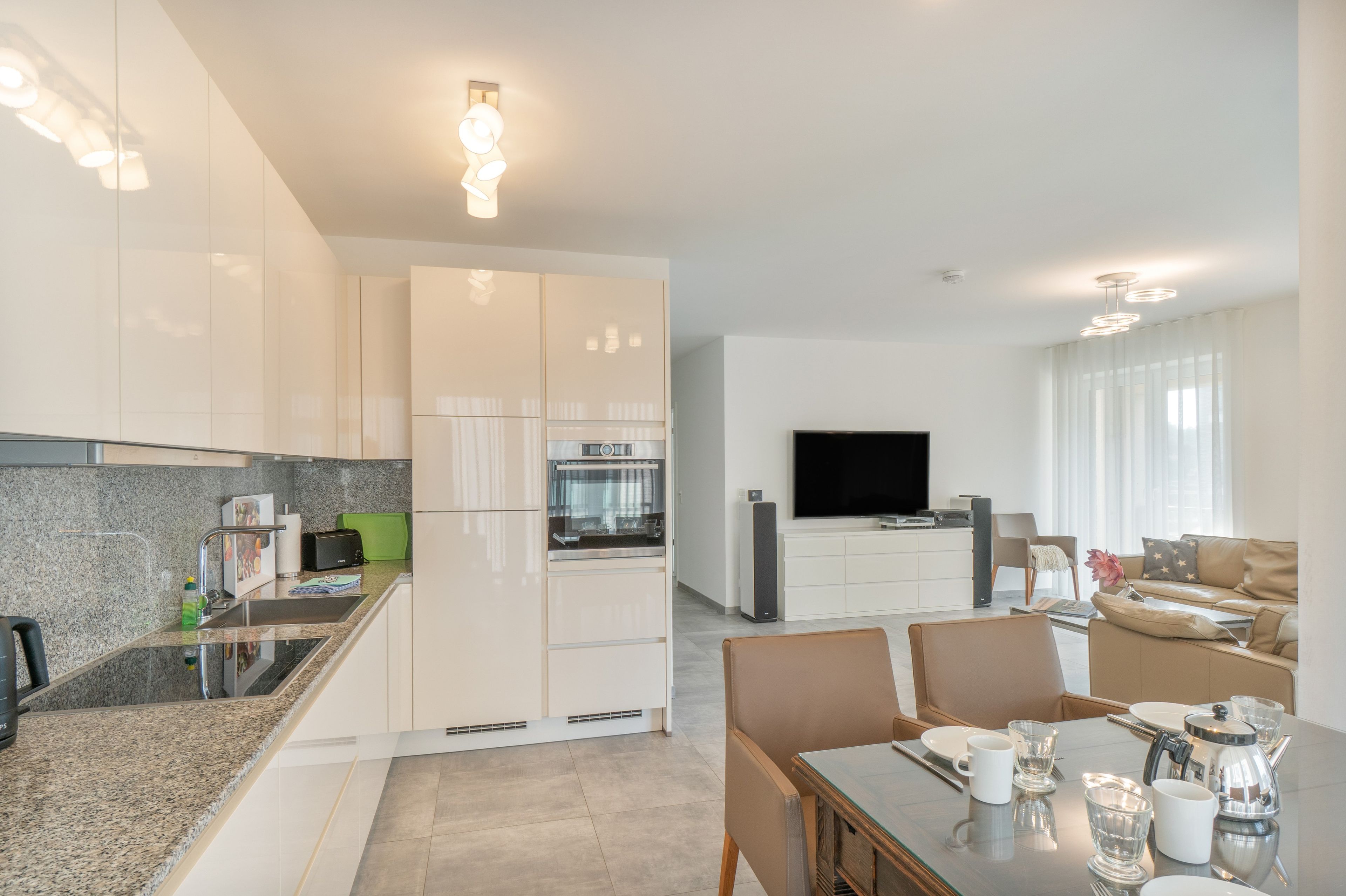 A modern kitchen with built-in appliances, a dining area with a table set for two, and a living room with a flat-screen TV and seating.
