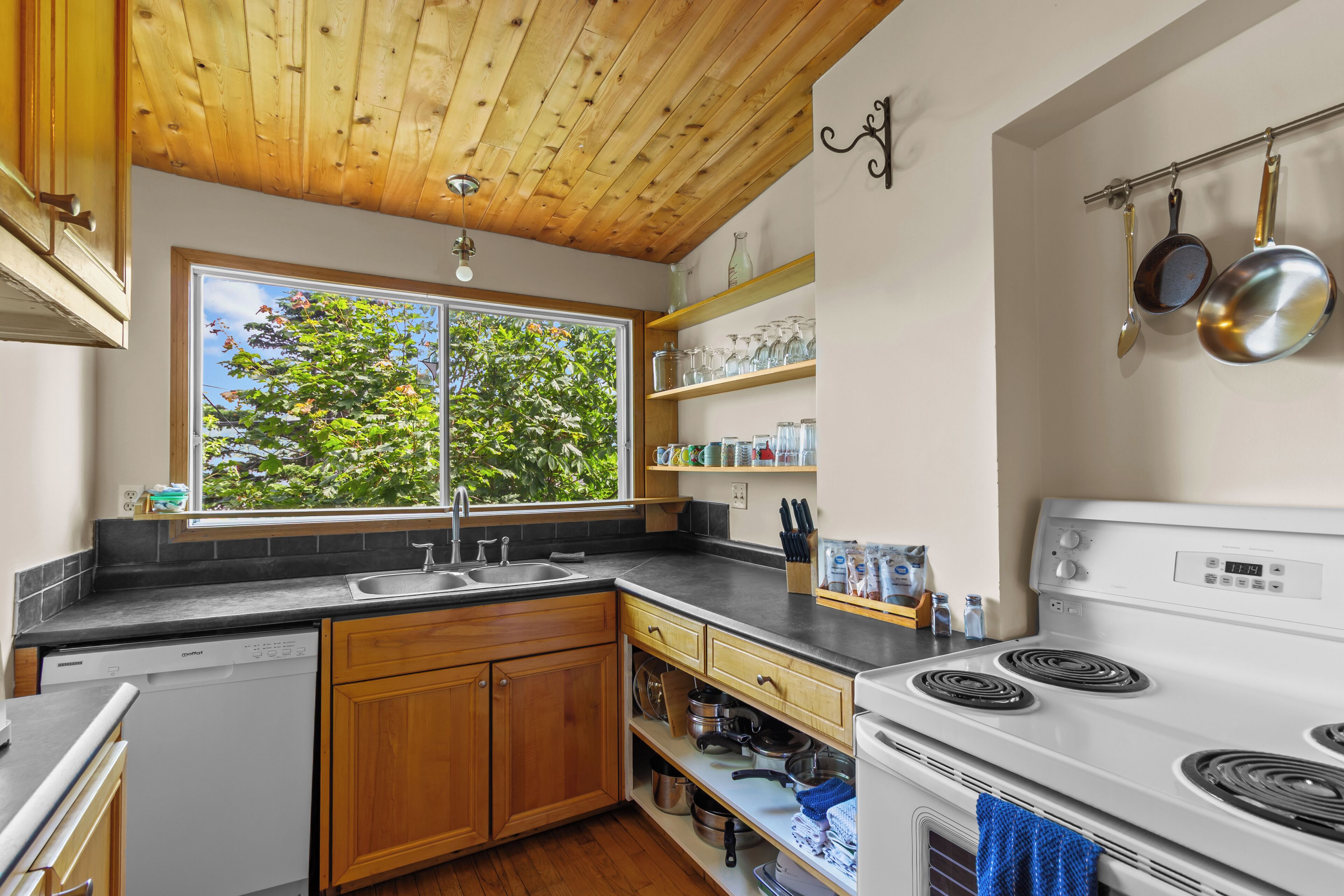 A kitchen with wooden cabinets, a double sink, a stove, and a window with a view of trees.
