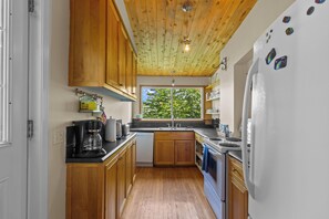 A kitchen with wooden cabinets, a black countertop, a window with a view of greenery, and a refrigerator with magnets. - Grand Heritage Home on Gower | 4BR (St. John's)