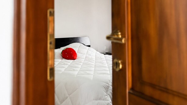 Un dormitorio con una colcha blanca, un objeto rojo sobre la cama y una puerta de madera.