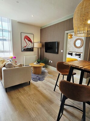 A modern living room with a sofa, a round coffee table, a dining table with chairs, a television mounted on the wall, and a patterned wallpaper. - Pelican House No.10 | Luxury Apartment | Walk to The Beach | Bournemouth Centre (Bournemouth, Christchurch and Poole)