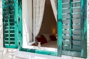 Ein Zimmer mit einem Bett, einem Fenster mit grünen Lamellen und einem weißen Vorhang.
