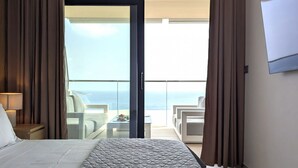 Habitación de hotel con cama, mesita de noche, lámpara, vista al mar a través de una puerta balcón y televisor montado en la pared.