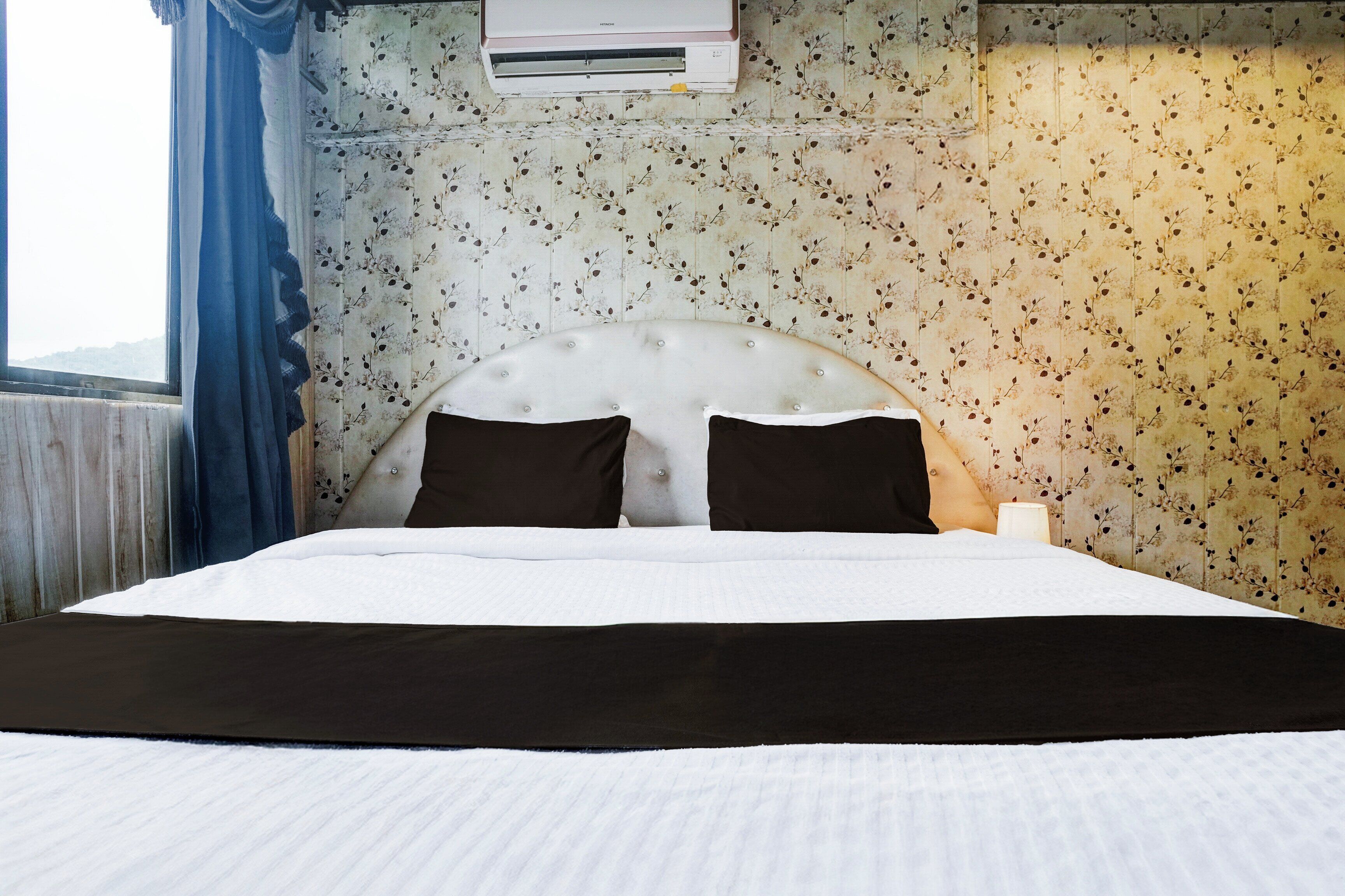 Habitación de hotel con cama, ventana con cortinas, aire acondicionado de pared y papel tapiz floral.