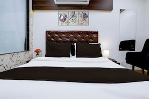 A hotel room with a large bed, a headboard, two pillows, a nightstand, a lamp, a chair, and a wall-mounted air conditioner.